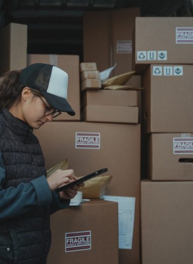 A warehouse employee checks inventory on a tablet beside stacked boxes marked fragile.